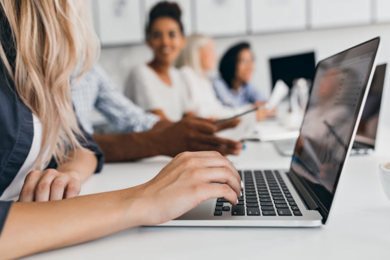 Blonde woman with elegant hairstyle typing text on keyboard in office. Indoor portrait of international employees with secretary using laptop on foreground.